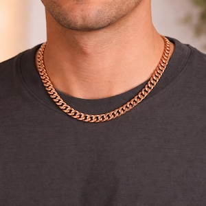 May include: A rose gold curb chain necklace. The necklace is made of interlocking links and is worn around the neck. The chain is a warm, metallic color and is displayed against a dark gray shirt.