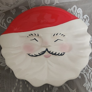May include: White ceramic Santa Claus dish with a red hat and a closed-eye smiling face. The dish has a scalloped edge.