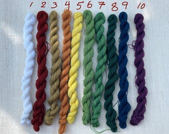 Puede incluir: Diez madejas de hilo de bordar en una variedad de colores, desde blanco hasta morado oscuro. Las madejas están numeradas y dispuestas en fila. El hilo es probablemente de algodón o fibra similar, adecuado para proyectos de bordado.