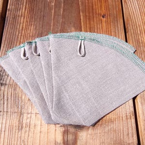 May include: A stack of five gray fabric coffee filters with green stitching along the edges and small loops for hanging. The filters are arranged on a wooden surface.