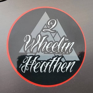 Puede incluir: Una pegatina negra y blanca con un borde rojo. La pegatina presenta un diseño de montaña estilizado con el texto "2 Wheelin' Heathen" en una fuente cursiva blanca.