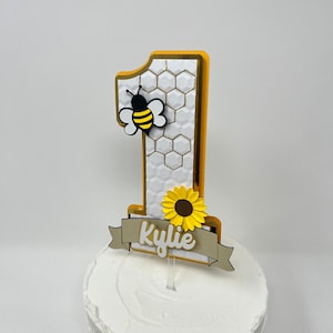 May include: A white cake with a yellow and gold number one cake topper. The number one has a honeycomb pattern and a small bee on it. A sunflower and a banner with the name "Kylie" are also on the topper.