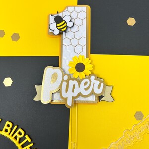 May include: A yellow and black bee with a white honeycomb number one cake topper. The number one is decorated with a sunflower and the name "Piper" is written in white with gold glitter on a white banner.