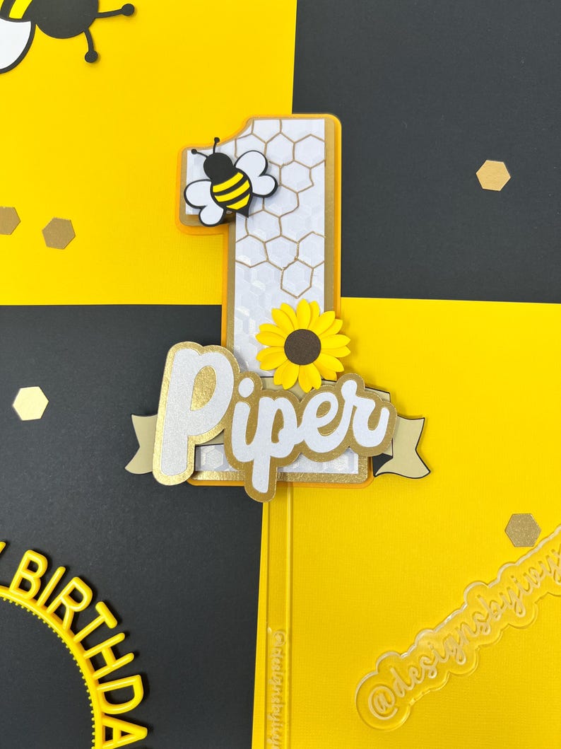 May include: A yellow and black bee with a white honeycomb number one cake topper. The number one is decorated with a sunflower and the name "Piper" is written in white with gold glitter on a white banner.