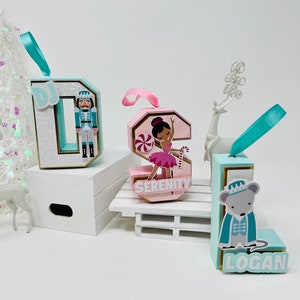 May include: Three personalized Christmas ornaments shaped like letters. The letter D is light blue with a gold trim and features a Nutcracker. The letter S is pink with a gold trim and features a ballerina. The letter L is light blue with a gold trim and features a mouse in a crown. The ornaments are decorated with glitter and have a ribbon for hanging.