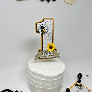 May include: A white cake with a yellow and black bee-themed cake topper. The topper features a number one, a bee, a sunflower, and a banner with the name "Kylie".