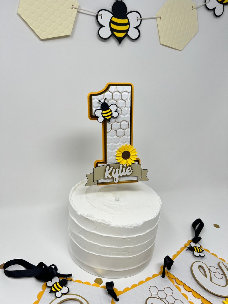 May include: A white cake with a yellow and black bee-themed cake topper. The topper features a number one, a bee, a sunflower, and a banner with the name "Kylie".