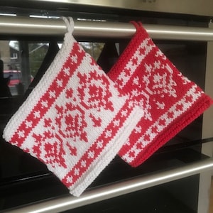 May include: Two red and white knitted pot holders with a geometric pattern hanging on a silver oven door handle.