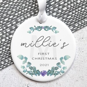 Peut inclure: Ornement en céramique blanche avec une couronne d'eucalyptus verte et des fleurs violettes. L'ornement dit "Millie's First Christmas 2021".