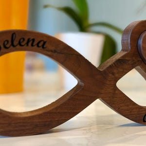 May include: A wooden infinity symbol with a heart-shaped accent. The infinity symbol is engraved with the names "Selena" and "Avery", and the heart has the word "Plumeria" engraved on it. The wood has a warm, brown tone.