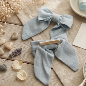 Puede incluir: Dos lazos para el pelo de tela azul claro con colas largas, uno con una pinza dorada. Los lazos están hechos de un material texturizado y están colocados sobre una superficie de madera con elementos decorativos.