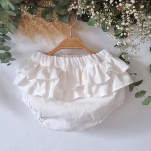 Puede incluir: Bombachos de beb&eacute; de lino blanco con volantes con un dise&ntilde;o de volantes superpuestos. Los bombachos est&aacute;n colgados de una percha de madera.