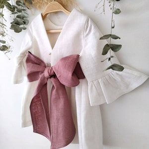 Peut inclure: Robe en lin blanc avec un grand nœud rose poudré. La robe a un col en V et des manches à volants. Elle est suspendue à un cintre en bois.