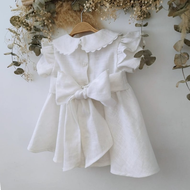 Puede incluir: Vestido blanco de lino con un gran lazo en la espalda y un cuello festoneado.