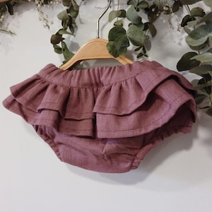 Puede incluir: Bragas de bebé de color rosa empolvado con volantes en capas. Las bragas están hechas de una tela texturizada y cuelgan de una percha de madera. Se ve vegetación en el fondo.