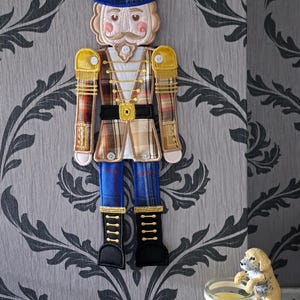 May include: A fabric nutcracker decoration with a blue hat, plaid jacket, and black boots. The ornament hangs against a grey and black patterned wallpaper. A candle with a dog figurine is in the foreground.