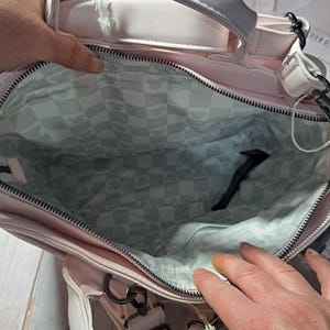May include: A light pink handbag with a top handle and a detachable shoulder strap. The interior lining features a checkered pattern with a repeating design. The bag has a zipper closure and metal hardware.