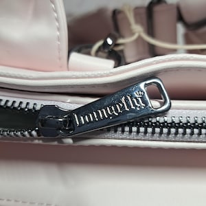 May include: Close-up of a light pink handbag with a silver zipper. The zipper pull has the word "Imunirella" engraved on it. The bag appears to be made of a soft material.
