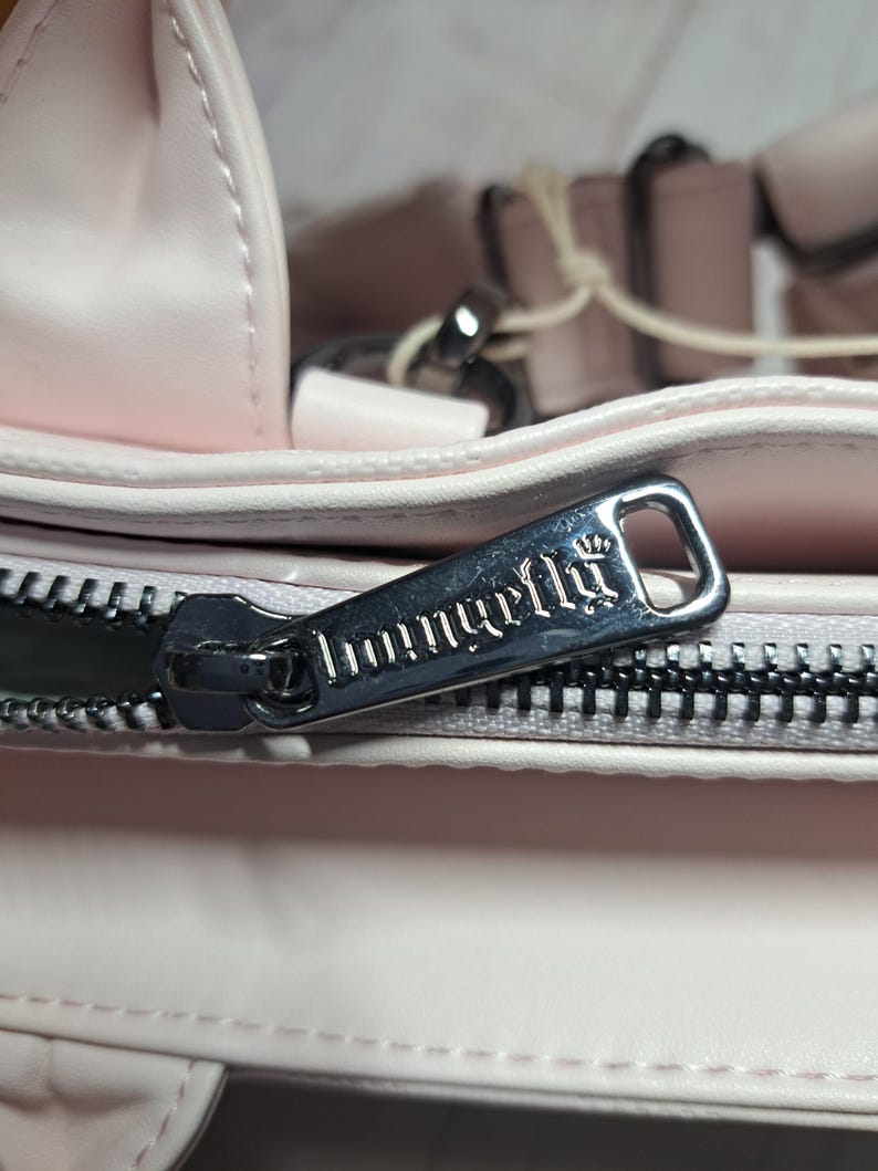May include: Close-up of a light pink handbag with a silver zipper. The zipper pull has the word "Imunirella" engraved on it. The bag appears to be made of a soft material.