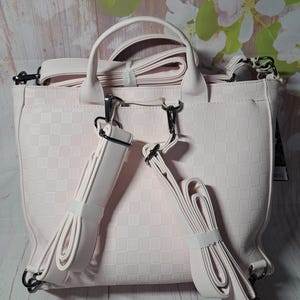May include: A light pink handbag with a checkered pattern. The bag has a top handle and adjustable shoulder straps. The straps are secured with black metal clasps. The bag is made of a soft material.