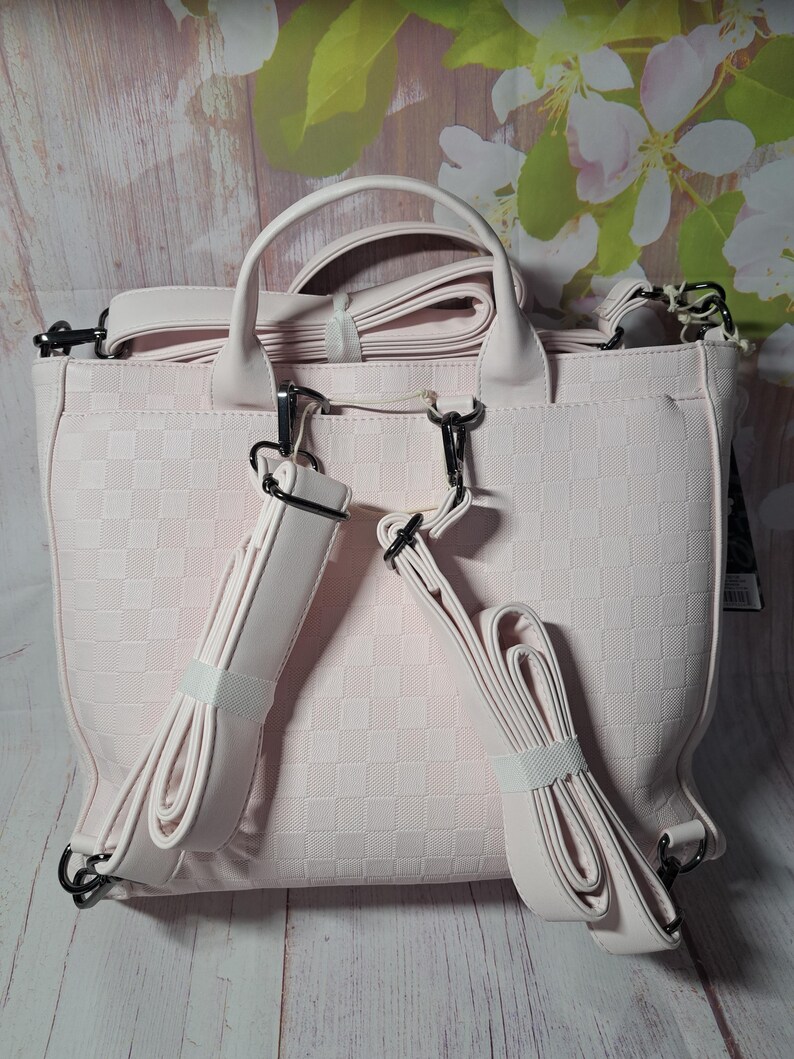 May include: A light pink handbag with a checkered pattern. The bag has a top handle and adjustable shoulder straps. The straps are secured with black metal clasps. The bag is made of a soft material.