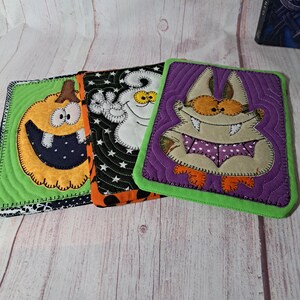 May include: Three Halloween-themed square coasters. Each coaster features a different design: a smiling pumpkin, a ghost, and a bat-like creature. The coasters have quilted details and are outlined in green, orange, and black.