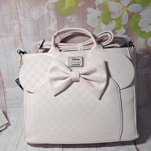 May include: A light pink handbag with a bow and mouse ears design. The bag has a checkered pattern and a Disney Loungefly logo. It features a top handle and a detachable shoulder strap.