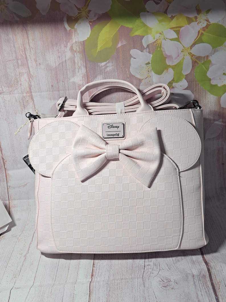 May include: A light pink handbag with a bow and mouse ears design. The bag has a checkered pattern and a Disney Loungefly logo. It features a top handle and a detachable shoulder strap.