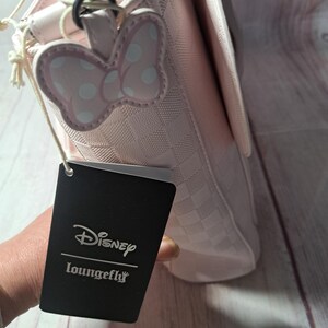 May include: A pink handbag with a checkered pattern and a Minnie Mouse bow-shaped charm. The bag has a flap closure and a silver-tone chain. A black tag with the Disney Loungefly logo is attached.