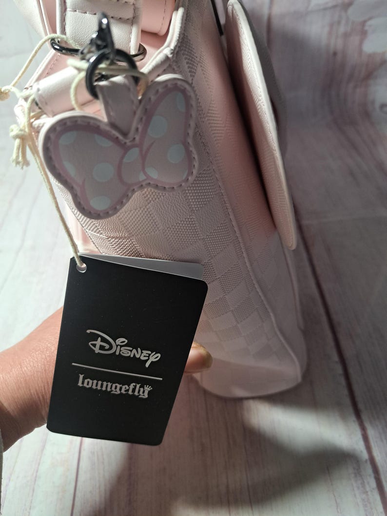 May include: A pink handbag with a checkered pattern and a Minnie Mouse bow-shaped charm. The bag has a flap closure and a silver-tone chain. A black tag with the Disney Loungefly logo is attached.