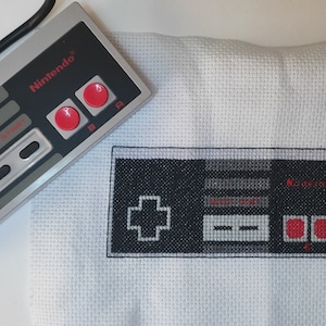 May include: A grey and black Nintendo controller beside a cross-stitch of the same controller. The controller has red buttons and the word "Nintendo" printed in red. The cross-stitch is black, grey, and red on a white background.
