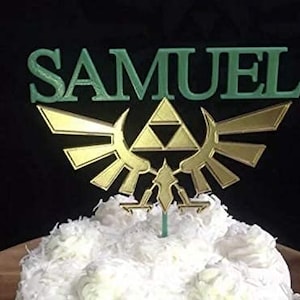 Puede incluir: Un adorno para pastel con el nombre "SAMUEL" en letras verdes sobre un emblema dorado. El emblema presenta un triángulo y alas estilizadas. El adorno está colocado sobre un pastel glaseado blanco, sobre un fondo negro.