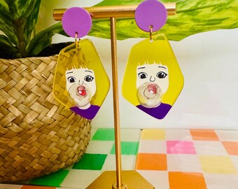 May include: A pair of yellow resin earrings with a cartoon image of a girl blowing a bubble. The earrings have purple accents and are displayed on a gold stand.
