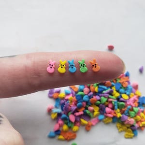 May include: Five colorful bunny shaped sprinkles with black eyes. The sprinkles are arranged in a row on a finger. A pile of colorful bunny shaped sprinkles is in the background.