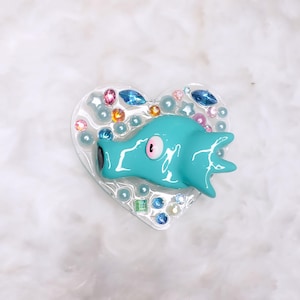 May include: A turquoise blue heart-shaped resin charm with a single eye and a textured surface. The charm is surrounded by white pearls and colorful rhinestones.