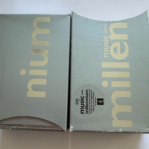 Peut inclure: Deux boîtiers de CD argentés. L'un porte le mot "nium", l'autre "music of the millennium". Le texte "Double cassette" et le chiffre "4" sont également visibles. Les boîtiers sont légèrement incurvés.