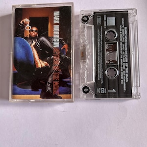 Puede incluir: Una cinta de casete con una foto en blanco y negro de un hombre con una chaqueta de cuero negra y pantalones vaqueros. La cinta de casete está etiquetada como "Mark Morrison Return of the Mack".