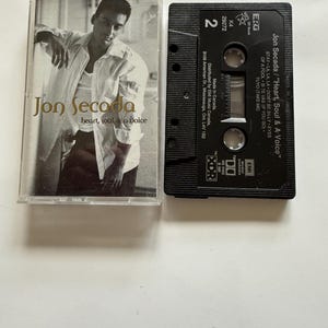 May include: A cassette tape and its case featuring Jon Secada's album "Heart, Soul & A Voice." The case has a black and white photo of a man. The cassette tape is black with white text.