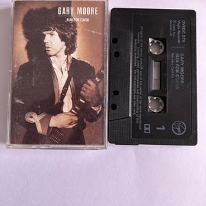 以下が含まれることがあります： 白いラベルが付いた黒いカセットテープ。ラベルには「GARY MOORE RUN FOR COVER」と「OVEDC 274」の文字が書かれている。テープは透明なプラスチックケースに入っていて、前面にゲイリー・ムーアの画像が印刷されている。