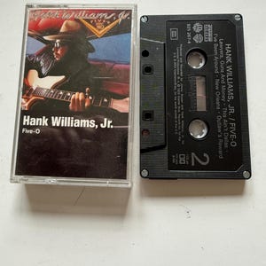May include: A black cassette tape with the title "Hank Williams, Jr. / Five-O" and the song titles "Lawyers, Guns And Money", "This Ain't Dallas", "I've Been Around", "New Orleans", and "Outlaw's Reward".