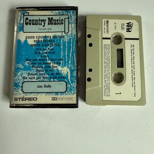 May include: A vintage cassette tape labeled "Country Music Volume One" with a blue and white cover featuring song titles and the artist "Jan Holly." The cassette is next to its open case, showing the tape reel.