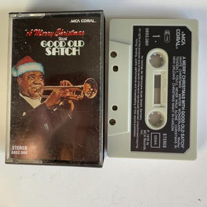 May include: A vintage cassette tape with a brown and white label. The label features a photo of a man in a Santa hat playing a trumpet. The text on the label reads "A Merry Christmas with Good Old Satch".