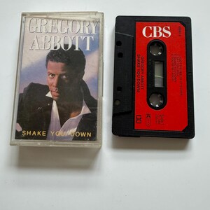 Puede incluir: Una cinta de cassette vintage del álbum "Shake You Down" de Gregory Abbott. La caja del cassette presenta un retrato del artista y el título del álbum. El cassette negro tiene una etiqueta roja con el logotipo de CBS y la lista de canciones.