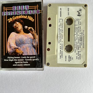 Puede incluir: Una cinta de casete blanca con una etiqueta negra. La etiqueta tiene el texto "ELLA FITZGERALD 20 Greatest Hits" y una foto de Ella Fitzgerald cantando en un micrófono. La cinta de casete está etiquetada como "Lado Uno" y enumera las canciones de la cinta. El texto "MP 666004" está impreso en el lado derecho de la cinta de casete.