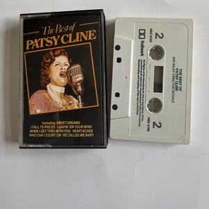 Può includere: Una musicassetta intitolata The Best of Patsy Cline con una custodia nera che presenta un ritratto della cantante e un elenco di brani tra cui Sweet Dreams e I Fall to Pieces. Il guscio bianco della cassetta è etichettato lato 2 con il marchio Hallmark.