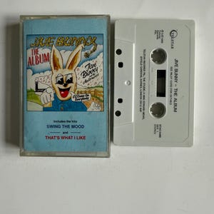 Puede incluir: Un casete vintage de "Jive Bunny - The Album" con una portada azul claro con un conejo de dibujos animados. El casete es blanco con texto. Incluye los éxitos "Swing the Mood" y "That's What I Like".