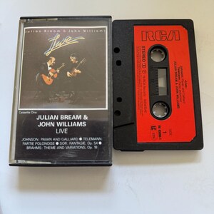 Puede incluir: Un casete vintage con la actuación en vivo de Julian Bream & John Williams. La caja del casete muestra a los músicos tocando la guitarra. El casete rojo tiene el logotipo de RCA y el título del álbum. La lista de canciones incluye obras de Johnson, Telemann y Brahms.