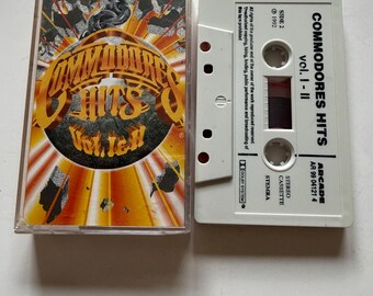 Może przedstawiać: Biała kaseta magnetofonowa z nadrukiem "Commodores Hits Vol. I - II" i "Arcade AR 99 04121 4" na etykiecie. Kaseta magnetofonowa znajduje się w przezroczystym plastikowym pudełku z kolorowym obrazkiem globusa z nadrukiem "Commodores Hits Vol. I" na nim.