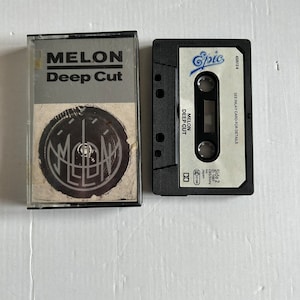 May include: A black cassette tape with a white label. The label has the text "MELON Deep Cut" and a logo. The cassette tape is in a plastic case with a black and white image of the band's logo.