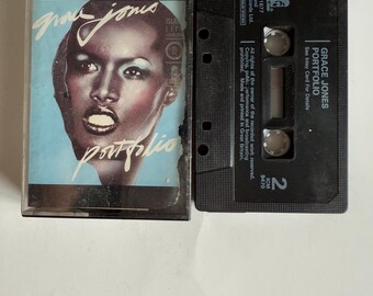 May include: A vintage cassette tape of Grace Jones' album "Portfolio." The cassette case features a portrait of Grace Jones with the album title. The black cassette tape is next to the case, with the album title printed on it.